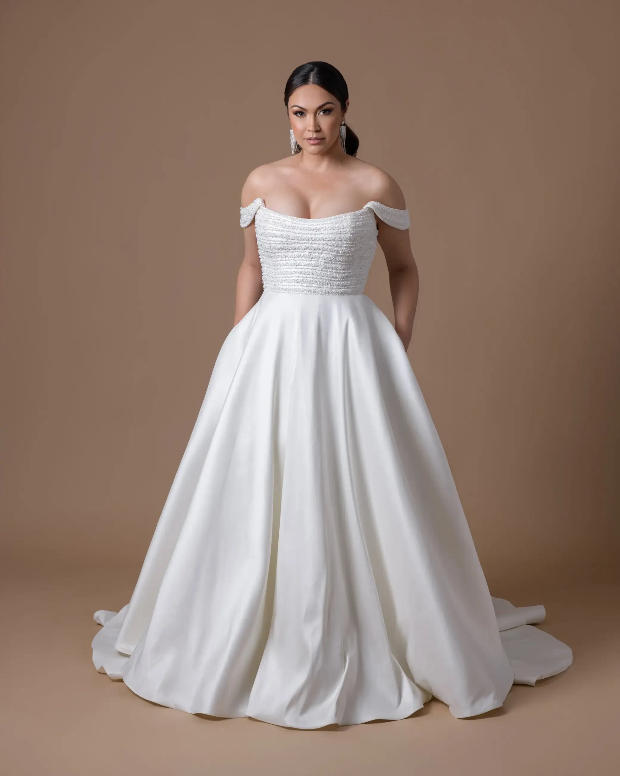 A woman stands confidently in a white, off-the-shoulder wedding gown with a beaded bodice and flowing skirt. The background is a soft brown, creating a serene ambiance.