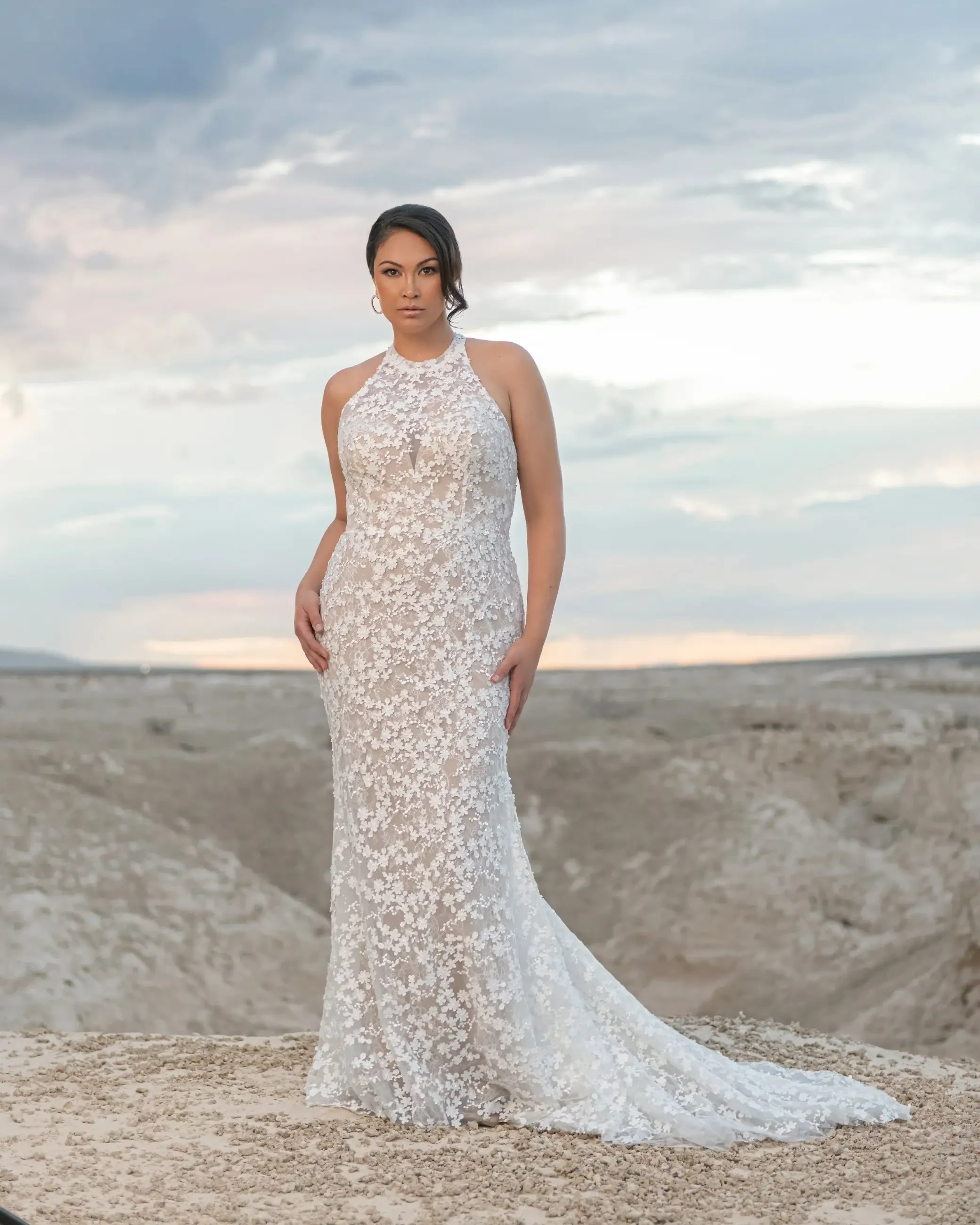 A woman in a floral lace gown stands confidently on a rocky desert terrain at sunset, exuding elegance and poise against a serene sky backdrop.